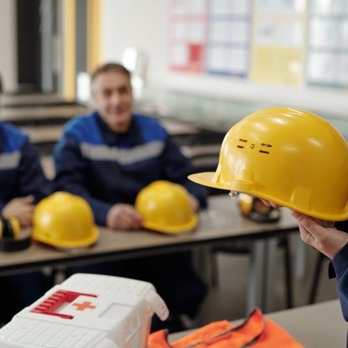 Young female worker in uniform holding protective helmet while standing in front of male colleagues at the lesson of basics of life safety